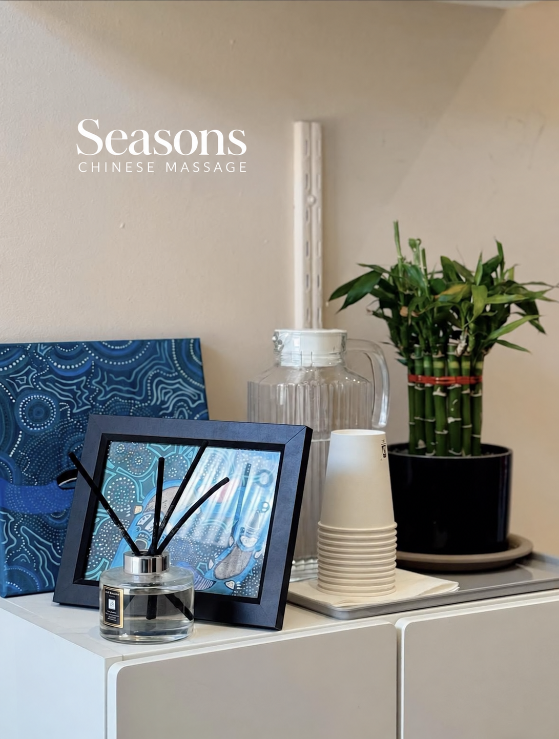 The Seasons Chinese Massage wordmark on the wall above a shelf with lucky bamboo and a reed diffuser